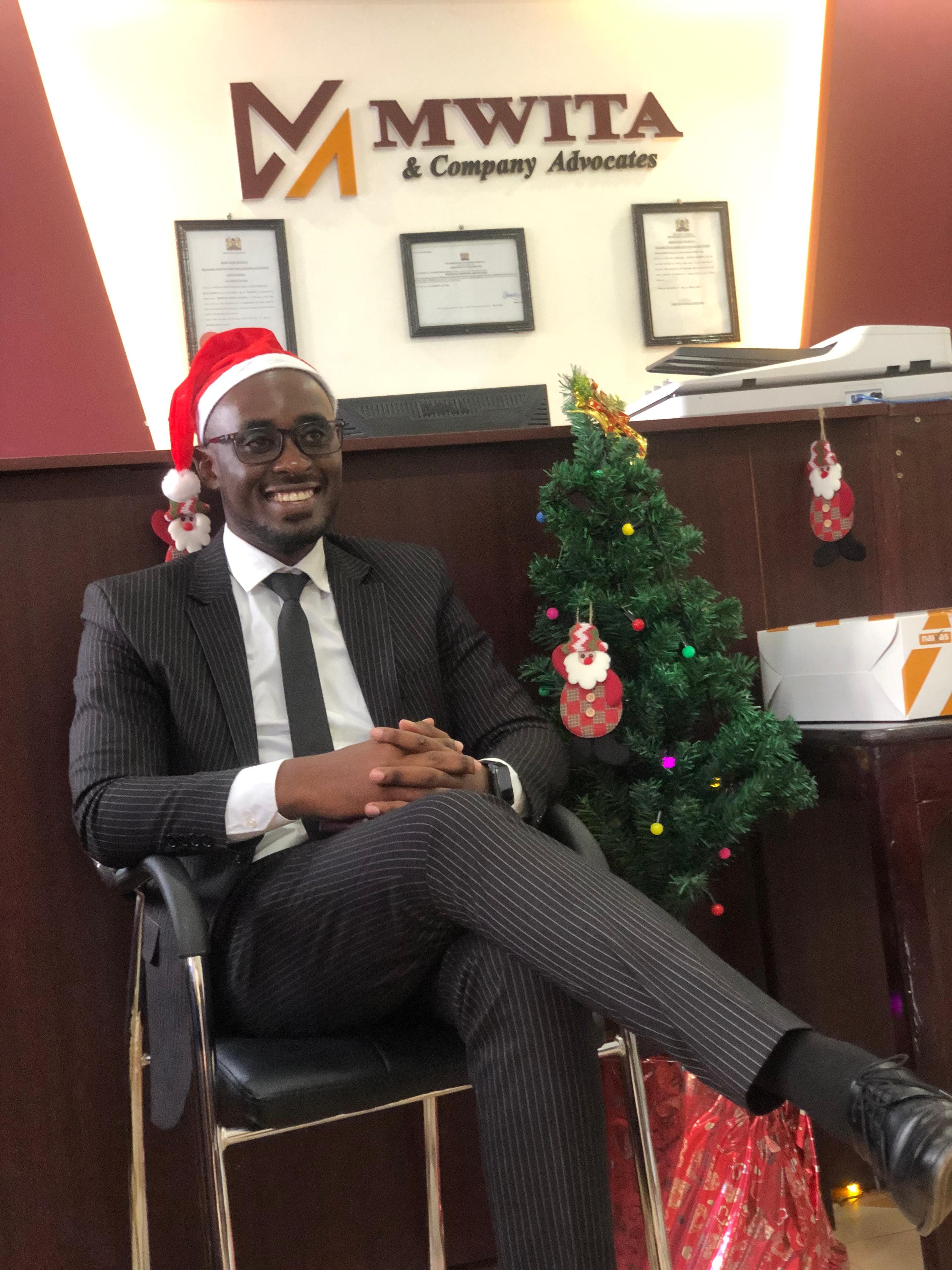 An advocate of Mwita & Co. in a pinstripe suit and Santa hat, seated by the office Christmas tree.