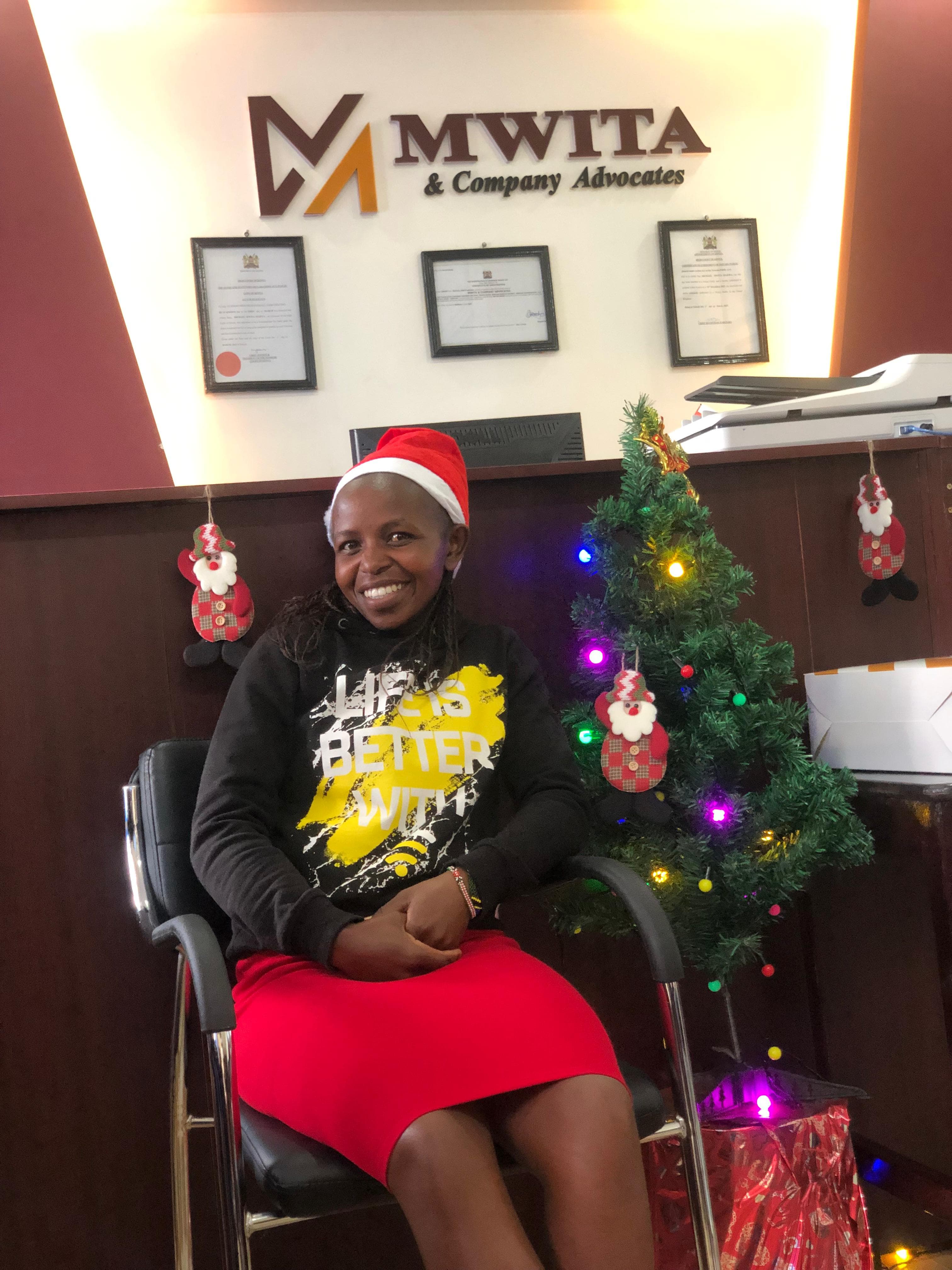 A smiling member of the Mwita & Co. team in a Santa hat beside a lit Christmas tree at the office.