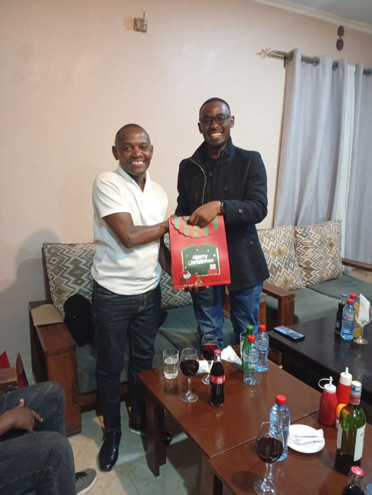 Two smiling men exchanging a festive Mwita & Co. gift bag in a lounge setting.