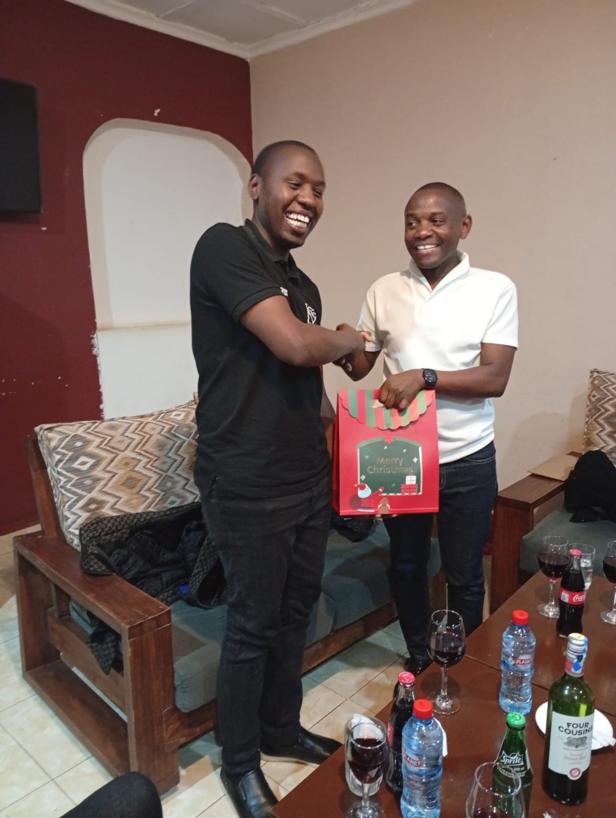 Two smiling members of the Mwita & Co. team exchanging a festive gift bag in a lounge setting.