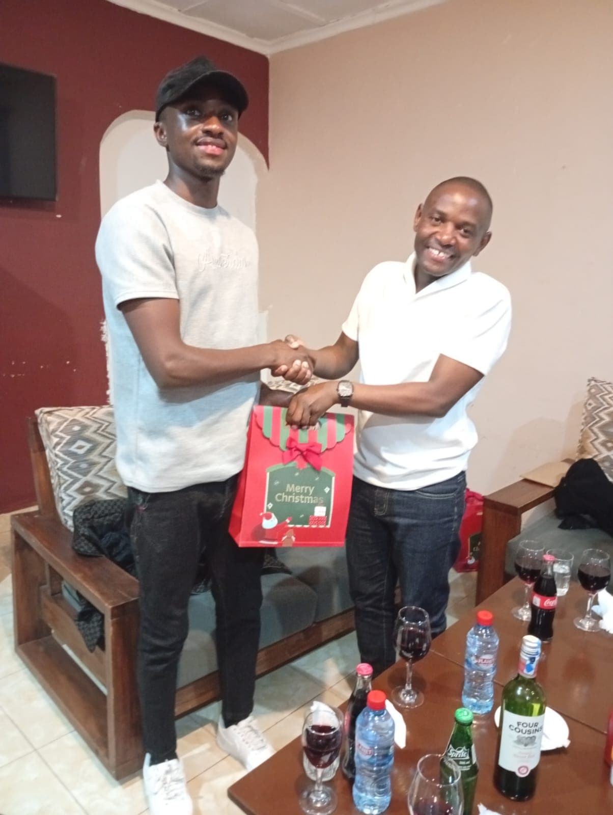 Two smiling people exchanging a festive Mwita & Co. gift bag in a lounge setting.